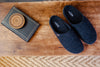 A pair of dark navy blue felt slip-on slippers placed on a rustic wooden table next to a decorative book with a wooden lid.