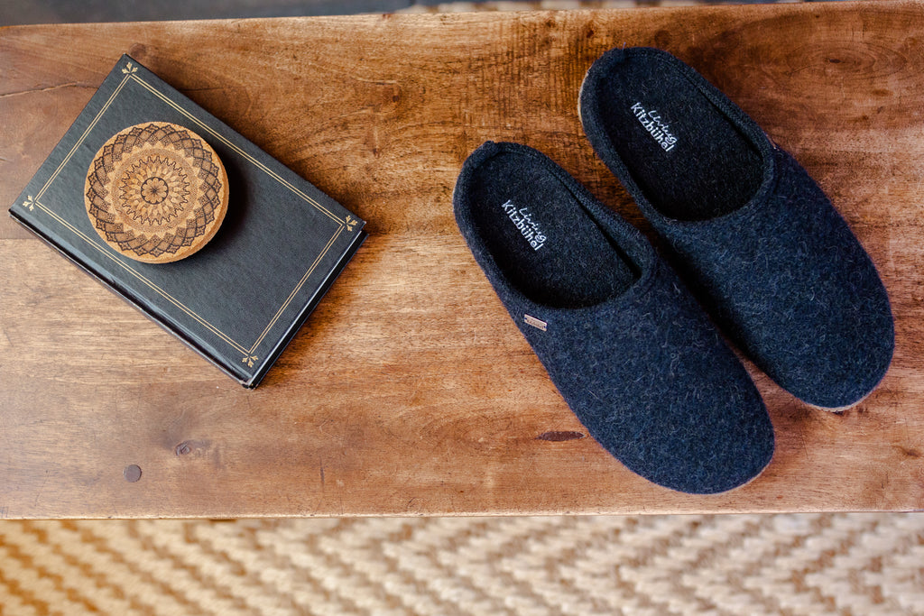 A pair of dark navy blue felt slip-on slippers placed on a rustic wooden table next to a decorative book with a wooden lid.