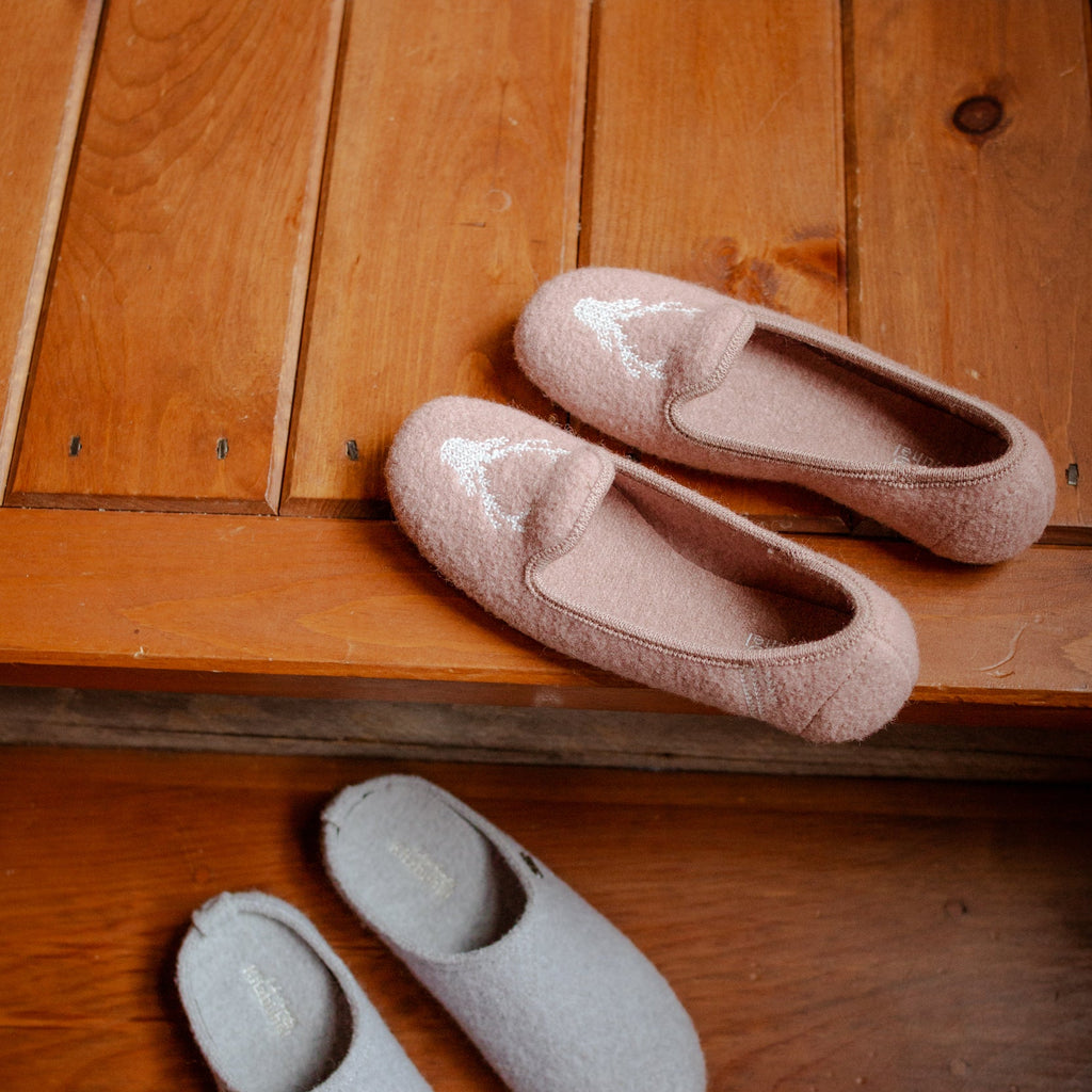 Two pairs of fuzzy slip-on slippers displayed on wooden steps: one pair in pale blush pink and one pair in light gray, with a patterned rug visible at the top edge.
