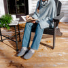A person sitting on a porch in a light blue knit cowl-neck sweater, blue jeans, and gray slip-on shoes, reading a book beside a small table with a plant and mug.