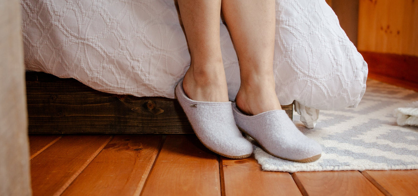 A person sitting on a bed wearing light gray knit slip-on slippers on a wooden floor, with white bedding and a patterned rug visible.