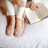 A person sitting on a white quilted bed wearing light pink knit slip-on slippers with white ribbed pants, with an open book resting beside them.