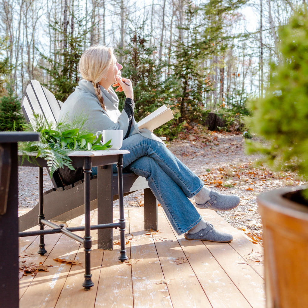 A woman sits in a wooden Adirondack chair on a wooden deck, reading a book outdoors with trees in the background.