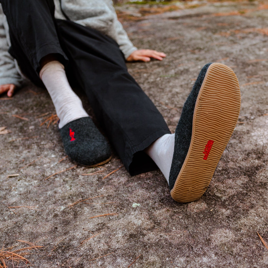 A person sitting outdoors on a textured stone surface, wearing dark gray wool felt slip-on shoes with white socks. The bottom sole of one shoe is raised, showing a tan rubber outsole and a small red logo.
