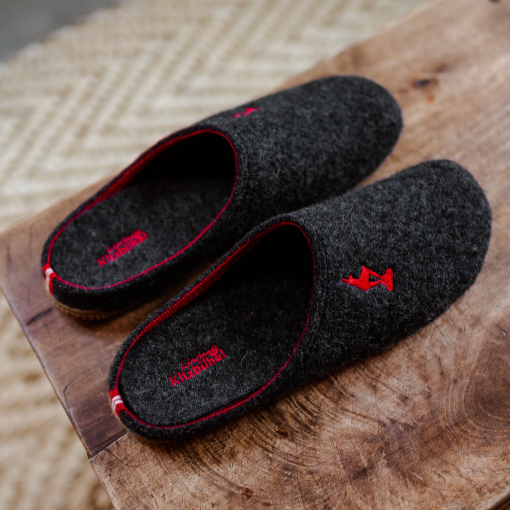 A pair of dark charcoal gray felt slippers with red trim and red embroidered details, displayed on a rustic wooden surface.