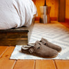 A pair of brown slip-on clog-style slippers with buckle straps sits on a small rug on a wooden floor in a cozy bedroom setting with a bed and lamp in the background.