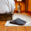 A pair of dark gray slip-on clog-style slippers with double straps, placed on a small rug on a wooden floor in a cozy bedroom setting.