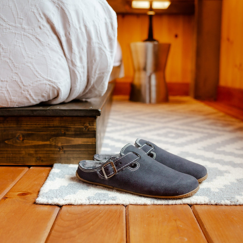 A pair of dark gray slip-on clog-style slippers with double straps, placed on a small rug on a wooden floor in a cozy bedroom setting.