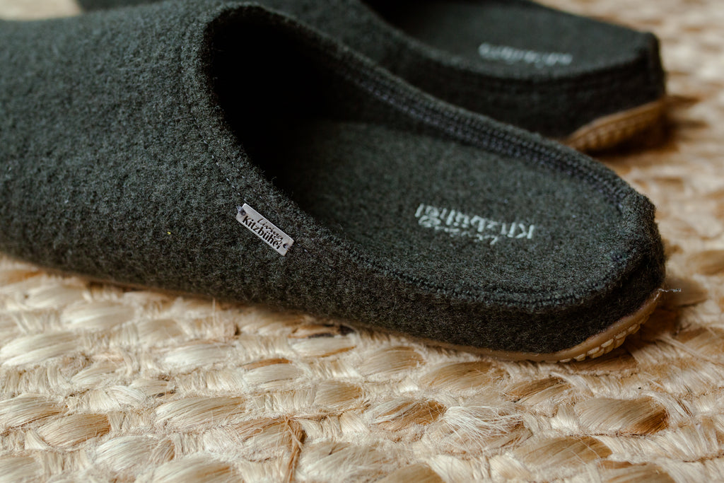 A close-up of a pair of dark charcoal gray wool slip-on slippers on a woven straw mat. The brand name 'KABIKUTU' is visible on the insole and a small side tag.