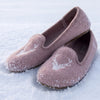 A pair of dusty pink felt slip-on slippers with white embroidered deer designs, resting on a bed of fresh snow.