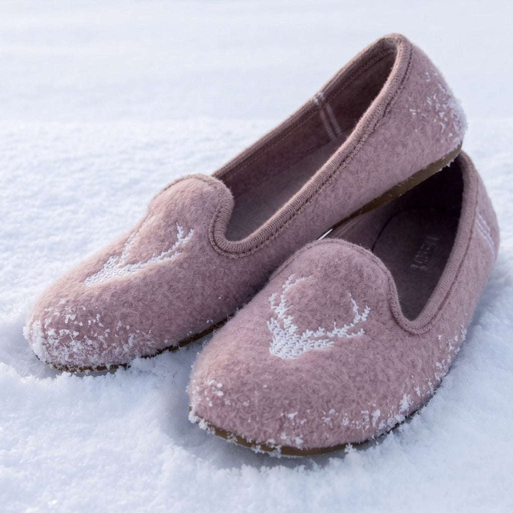 A pair of dusty pink felt slip-on slippers with white embroidered deer designs, resting on a bed of fresh snow.