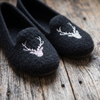 A pair of black felt slip-on slippers with white embroidered deer head motifs, displayed on a rustic weathered wood surface.